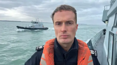 A uniformed officer, standing on the deck of a Border Force cutter, wearing a lifejacket and staring solemnly into camera. A second Border Force vessel is at sea in the background.