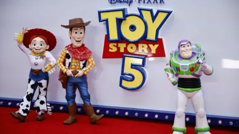 Toy Story characters Jessie, Woody and Buzz Lightyear stand in front of a sign for Toy Story 5