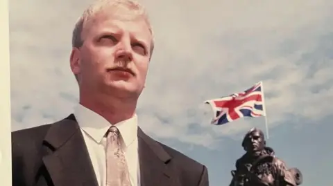 MOD In a photo from the early 1990s, a young man poses beside a statue of his image. The man has blond hair and moustache, and the statue in the background can be seen in military uniform with a union jack flag protruding from the backpack