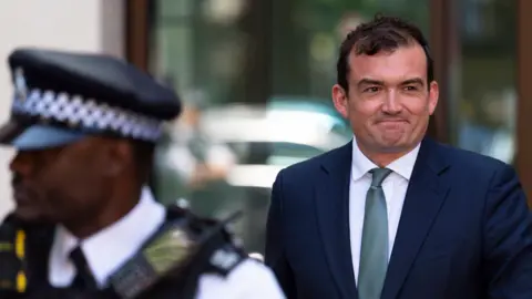 Patrick Spencer leaves a court, dressed in a blue suit and green tie. He is smiling at the cameras. In front of him is a police officer, dressed in uniform. 