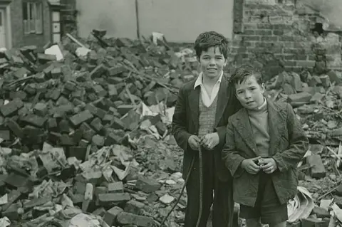 John Blakemore Two children stood in front of a collapsed building with a pile of bricks next to a damaged wall. They wear coats over jumpers and one appears to hold a length of rope.