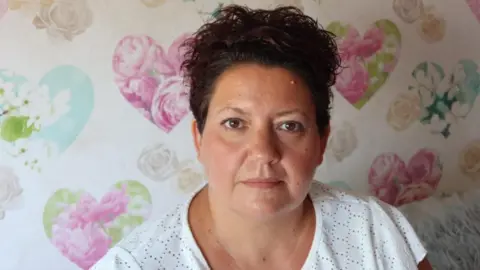 A woman with dark, curly hair tied back wearing a white broderie t-shirt in a room with cream and pastel coloured heart floral motif wallpaper.
