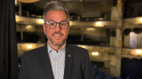 BBC/Richard Moss Jonathan Higgins is standing and smiling in the Tyne Theatre. He has grey hair, a beard and glasses and is wearing a dark suit jacket with a white shirt with a blue, pink and grey butterfly pattern.