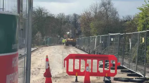 The photo is framed by metal construction fencing on either side. Down the centre we can see a road with its surface stripped away. Cones and red barriers are scattered about, and yellow construction vehicles can be seen in the distance, as well as workers in high vis clothing
