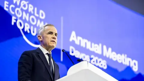Canada Prime Minister Mark Carney speaks during a plenary session in the Congress Hall. He is wearing a black suit and tie with a white button-up shirt underneath. He is standing behind a white podium, and is photographed from his lower right side. Behind him is a big blue screen with white lettering that reads: WORLD ECONOMIC FORUM, Annual Meeting Davos 2026