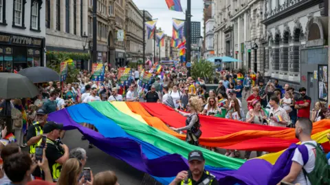 Matthew Horwood/Pride Cymru A crowd of people gather around a rainbow flag that surrounds a person with long hair wearing a dark top and tattoos on their arm.