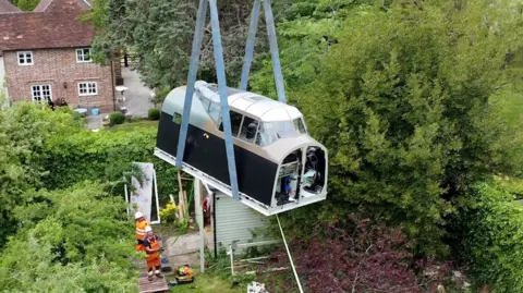 The cockpit being winched over the garden where it was built, with large cables holding it upright.