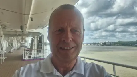 David Munster David taking a selfie on a ship deck smiling. He is near a railing and there's a large brown river behind him 