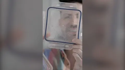 A man looks through the clear plastic lid of a food storage container. He's smiling and is wearing a multicoloured striped shirt.
