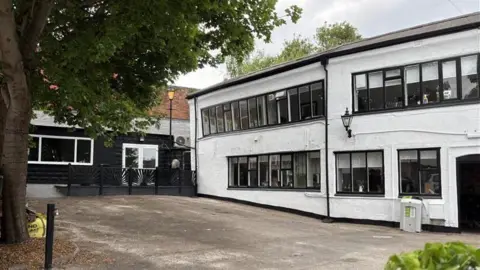 The Dynamo Project A white two-storey building with black windows. There is a big tree in front of the building and a small building to the left.