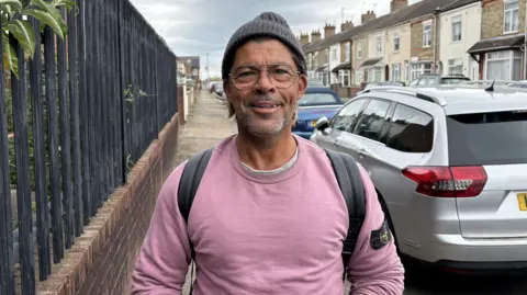 Ben Schofield/BBC Giuseppe Pizzo standing in a street, by metal railings and a car, with a pink jumper on, a grey hat, a beard and glasses. He is smiling at the camera. A row of houses are to the right. 