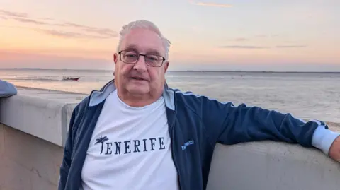 A man with short white hair, black rectangular glasses, a white top which says Tenerife and a navy jacket is leaning against a wall with the pink hues of sunrise behind him, reflecting softly on the water.