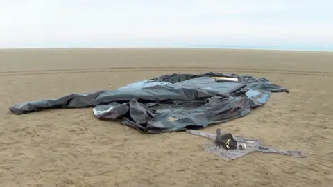 The remnants of an inflatable boat lies abandoned on a beach in northern France. A large amount of what looks like black plastic sheeting folded randomly on a large stretch of flat sand. 