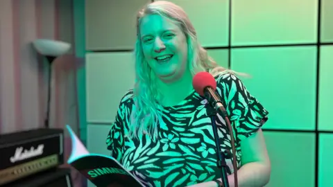 Poet Ellen Clayton standing in front of a microphone, smiling and holding her book of poetry.