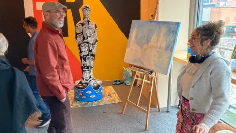 John Macauley Eric Cantona is talking to a female artist. They are stood in an art gallery with canvas' with paintings are being displayed. 