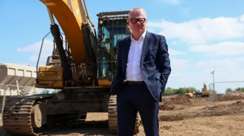 West Midlands Combined Authority A man in a dark suit and white shirt is standing in front of a digger looking at the camera. He is wearing glasses.