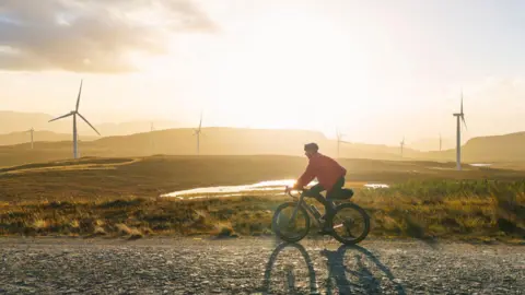 Getty Images A man in a red jacket rides a bike along a gravel road, past wind turbines as the sun sets in Scotland.