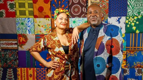 A woman and man in bright African-inspired clothing, standing in front of a multi-coloured quilted display. The woman is in a jacket which has brown, gold, black and white patterns, over a black crop top. She is wearing large silver dangly earrings and has a green, orange, yellow and red scarf knotted on her head. The man is dressed in a jacket with large blue, purple and orange circular patterns over a navy polo shirt. His head is shaved and he has a neat grey beard.