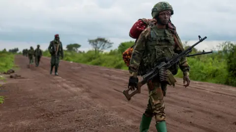 Reuters An M23 fighting in camouflage uniform walking along a dirt road in DR Congo, followed by his comrades. He is holding a gun, and carrying a large backpack.