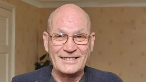 David Newton: A man is smiling at the camera in an image from 2016. He is bald, has some stubble and wears thin framed glasses. He is wearing a navy jumper, with a grey top underneath a has a hooped earring in his left ear. He is  inside a room with rust colour wallpaper that has red diamond shaped patterns. There is a white door behind him to his left and a green plant.