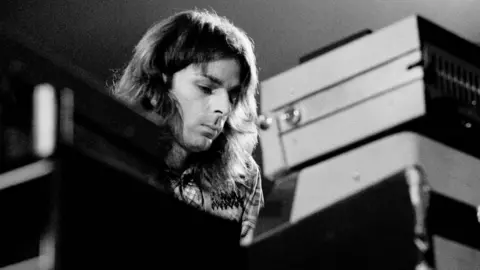 Getty Images A black and white photograph of Rick Wright performing, his face just visible over a stack of keyboards. He has shoulder length hair and a plaid shirt.