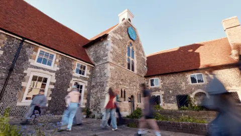 A stone building with a red tiled roof. A line of people are walking in front of it, but their motion is blurred by the camera.