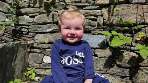 Smiling baby in a blue jumper sitting in front of a brick wall.