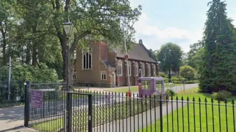 The outside of a school where its name can be seen on a purple plaque. A chapel is in the school grounds.