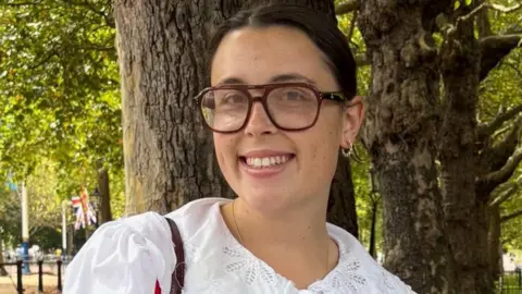 Isabel Mills in a white blouse standing outside in front of a tree.
