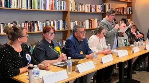 A long table with several people sat along it. Some are wearing political party rosettes. A man in the centre is talking with a microphone.