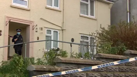 Street photograph of Judith Rhead's home on Market Street, Pembroke Dock. A police officer stands in the door way, wearing a face covering. The door frame is a peach colour, and there are five white windows on the yellow painted building. A support rail is at the forefront of the photograph, just behind a brick inclined wall. Blue and white police tape is seen across the bottom right hand corner. 