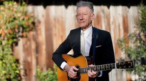 Celtic Connections Lyle Lovett, dressed in a black jacket, white shirt and off-white tie, stands in front of a fence and some shrubs, holding an acoustic guitar. 