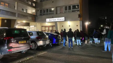 A crowd of people stood at the entrance of Barton House in Bristol. It is dark and they are all wearing coats and jackets, many with their hoods up.