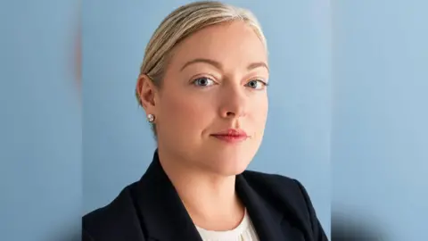 Victim support NI A blonde woman looking at the camera against a blue background. She is wearing a dark jacket 