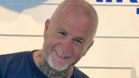 Simon Beston smiling at the camera. He has very short shaved grey hair and beard. He is wearing a blue t-shirt and has a neck tattoo. 
