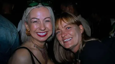 Aimi Rix with Dianne Cleary smile at the camera with their heads touching together. Ms Rix is wearing a necklace and black strappy top, she has white blonde hair, with sunglasses on her head, and is wearing red lipstick. Ms Clearly has blue eyes and light brown hair, cut with a fringe. Both women are wearing brown dangling earrings. 