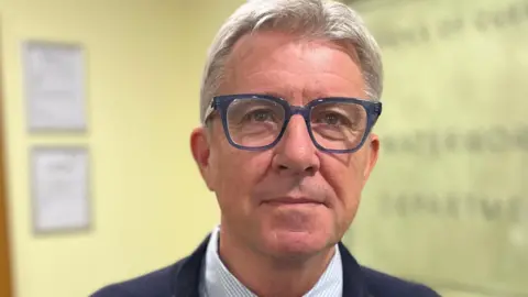 A photo of Deputy Mark Helyar who is a man with grey hair in a small quiff, wearing blue rimmed glasses. He is wearing a blue jacket, with a blue and white checked shirt, with a purple tie. 