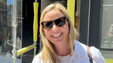 A woman with sun glasses on smiling at the camera. she has blonde hair and is wearing a white jacket and a white t-shirt