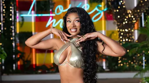Getty Images Megan Thee Stallion poses in a gold bikini with a halterneck design. Her hands are held under her chin, showing off her long, manicured nails. Fairy lights and neon signs can be seen in the background.