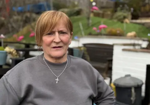 Diane Morgan looking at the camera. She has a blonde and brown fringe and is wearing a grey jumper with a silver necklace that has a cross pendant. She is sat in the garden.