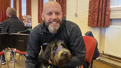 Simon Kelly has a beard and is wearing a black jacket. He's holding Pablo, a dark labradoodle.