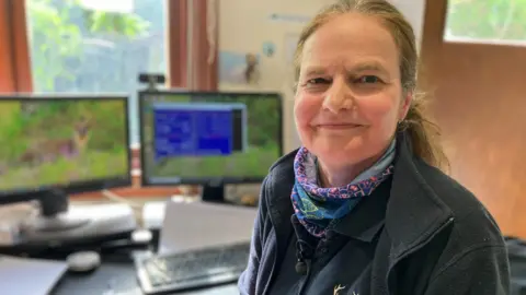 BBC / Fiona Irving A white woman with long, tied-up hair smiles at the camera. She is wearing a multi-coloured neck scarf and a dark fleece. In the background are two computer screens.