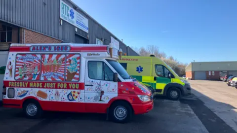 A red and white coloured ice cream van is parked outside a business unit. Parked alongside the van is an ambulance