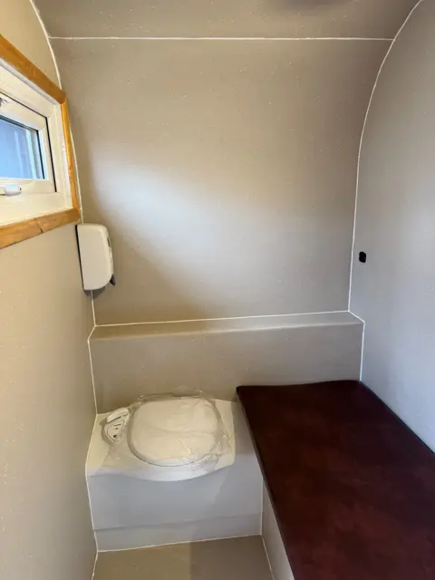 Stuart Johnson The interior of a sleeping pod. It is a small space with bare grey walls and a window. There is a white toilet covered with plastic, a soap dispenser and a bed with a purple mattress.