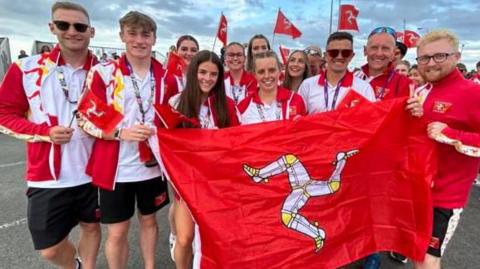 Athletes dressed in the Manx red and white kit smiling while holding up a red Manx flag