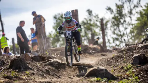 Jerry Tatton/ @jwdt.photography A young person is riding down a dusty incline while people watch on. The mountain biker is wearing a grey helmet and top with green goggles and yellow gloves. The number 9 is taped on to the front of their bike. They are riding over the stumps and roots of trees. There is greenery out of focus in the background.