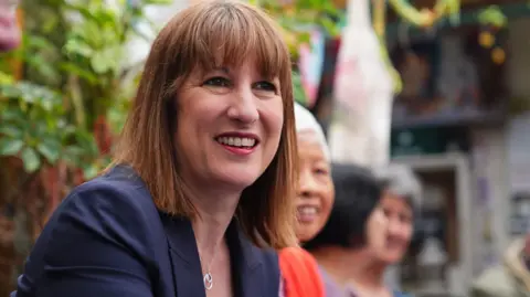 PA Media Rachel Reeves is smiling, with her eyes not looking directly at the camera. She is has brown hair with a fringe and is wearing a navy jacket and a necklace with two circles. There are people sitting alongside her but they are mostly blurred as she is the focus.