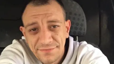 George Robertson looking straight at the camera. He is wearing a light grey hoodie. He is smiling and appeared to be sitting in a van.