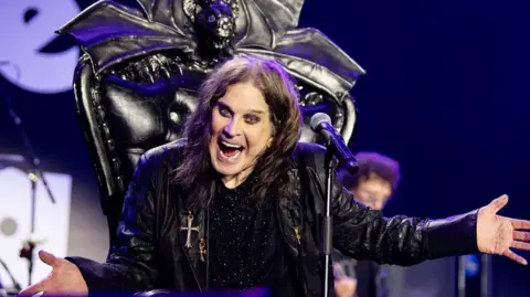 Ross Halfin Ozzy is sat in his throne on stage at the concert. He has his arms outstretched with the microphone in front of him. He is wearing all black with a bat shape on the back of his throne.