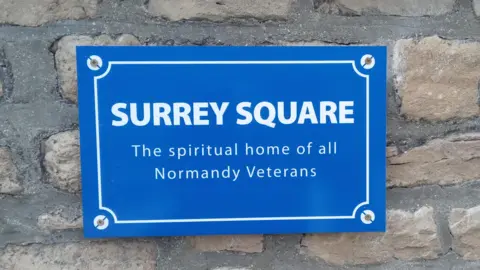 Ian Allen A blue plaque with white writing on a brick wall. The plaque reads: "SURREY SQUARE - The spiritual home of all Normandy Veterans".